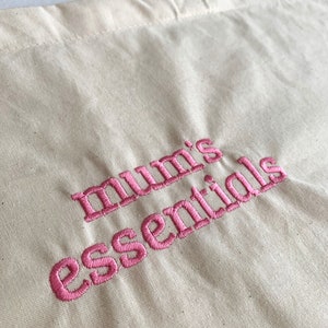 Puede incluir: Una bolsa de lona blanca con texto bordado en rosa que dice "mum's essentials" en una fuente de escritura cursiva. El texto est&aacute; en el centro inferior de la bolsa. La imagen est&aacute; etiquetada como "font 1" en la esquina inferior derecha.