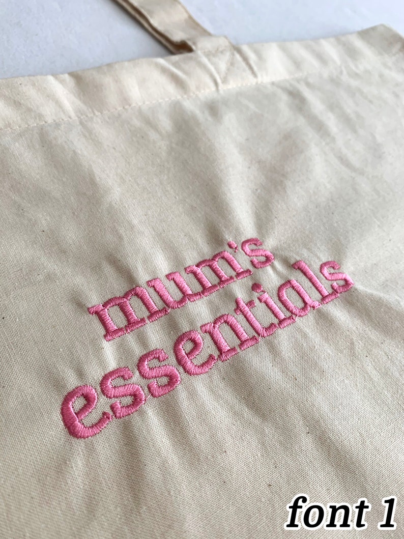 Puede incluir: Una bolsa de lona blanca con texto bordado en rosa que dice "mum's essentials" en una fuente de escritura cursiva. El texto est&aacute; en el centro inferior de la bolsa. La imagen est&aacute; etiquetada como "font 1" en la esquina inferior derecha.