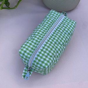Op de afbeelding: Een groen en wit geruit etui met een zilveren rits. De rechthoekige etui is gewatteerd en heeft een kleine lus aan de zijkant. De etui ligt op een roze ondergrond.