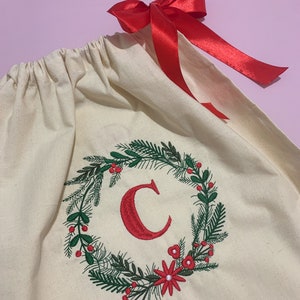 May include: A white canvas gift bag with a red ribbon. The bag is decorated with an embroidered wreath with green leaves, red berries, and red flowers. The letter "C" is embroidered in red in the center of the wreath.