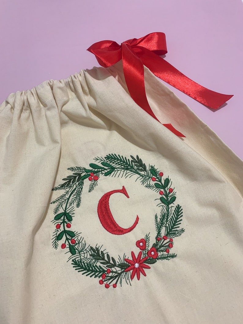 May include: A white canvas gift bag with a red ribbon. The bag is decorated with an embroidered wreath with green leaves, red berries, and red flowers. The letter "C" is embroidered in red in the center of the wreath.