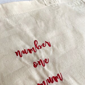 Puede incluir: Una bolsa de lona blanca con texto bordado en rojo que dice "number one mum" en una fuente cursiva. El texto est&aacute; en la fuente 3.