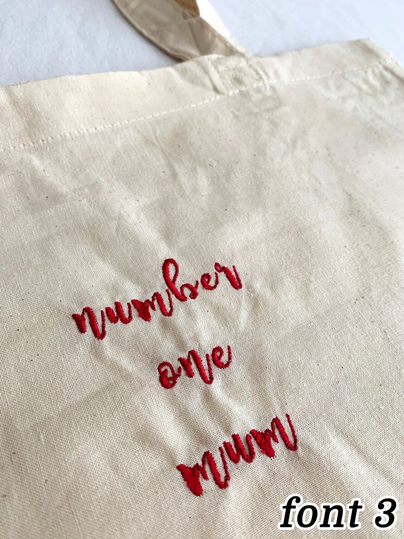 Puede incluir: Una bolsa de lona blanca con texto bordado en rojo que dice "number one mum" en una fuente cursiva. El texto est&aacute; en la fuente 3.