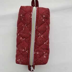 May include: A red quilted zippered pouch with a white dragonfly pattern. The pouch has a top handle and a zipper closure.