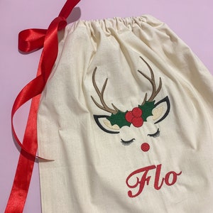 May include: A white canvas gift bag with a red ribbon. The bag has an embroidered reindeer face with a red nose and green holly. The name "Flo" is embroidered in red below the reindeer.