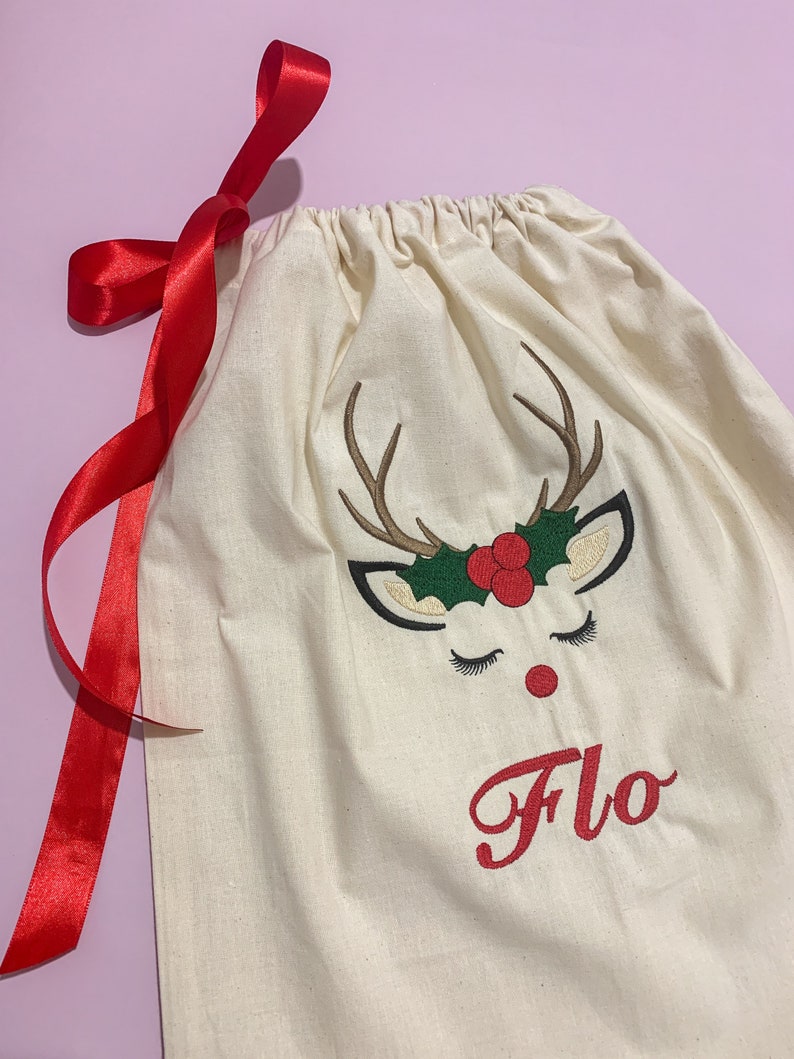 May include: A white canvas gift bag with a red ribbon. The bag has an embroidered reindeer face with a red nose and green holly. The name "Flo" is embroidered in red below the reindeer.