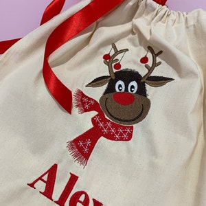 May include: A white canvas gift bag with a red ribbon, a reindeer design, and the name "Alex" embroidered in red.