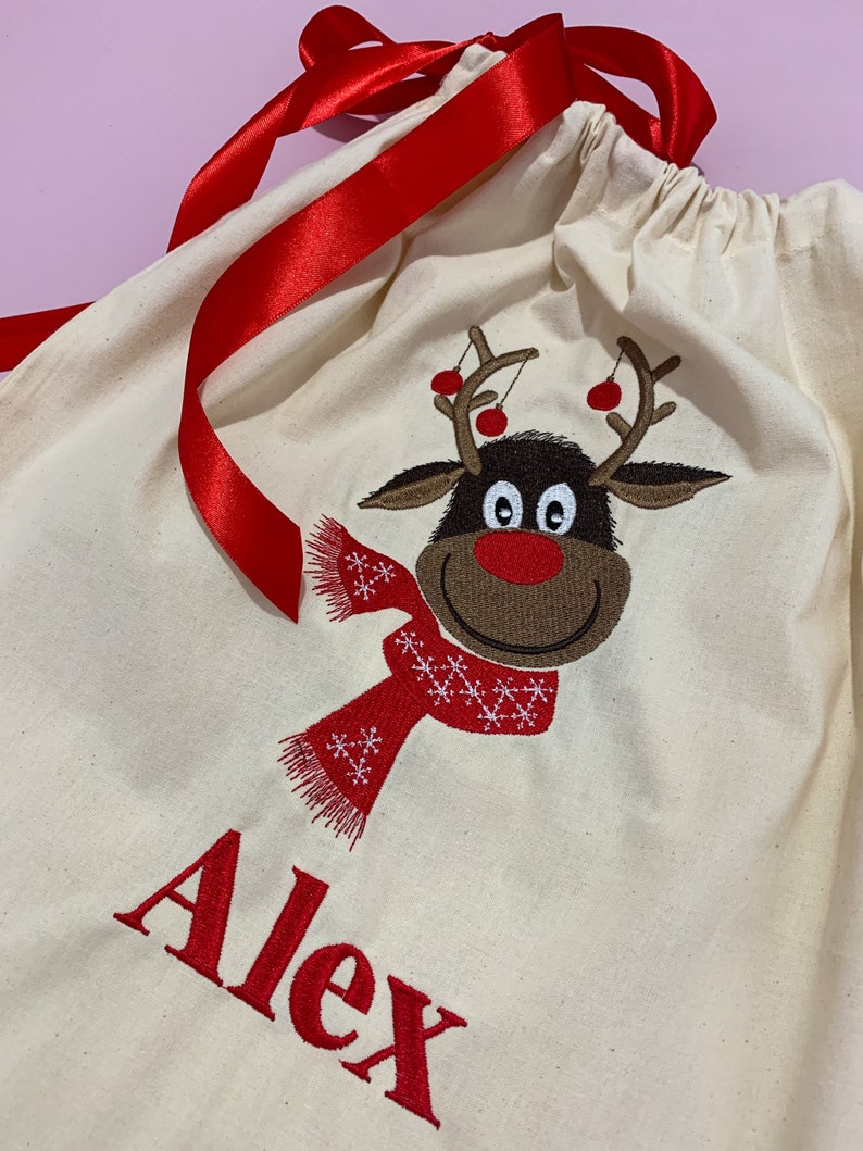 May include: A white canvas gift bag with a red ribbon, a reindeer design, and the name "Alex" embroidered in red.