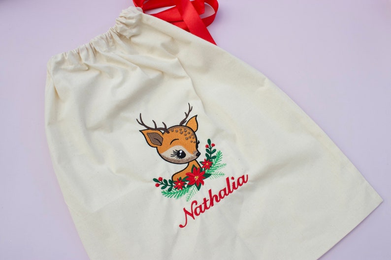 May include: A white canvas gift bag with a red ribbon. The bag has an embroidered design of a cartoon deer with a red bow and green holly. The name "Nathalia" is embroidered in red below the deer.