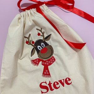 May include: A white canvas gift bag with a red ribbon. The bag features an embroidered reindeer with a red scarf and the name "Steve" in red.