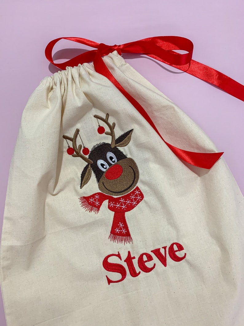 May include: A white canvas gift bag with a red ribbon. The bag features an embroidered reindeer with a red scarf and the name "Steve" in red.
