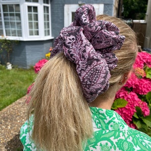 Puede incluir: Una gran scrunchie estampada en tonos morados, negros y blancos adorna una cola de caballo rubia. La scrunchie presenta un diseño intrincado. La persona lleva una camisa estampada verde y blanca.