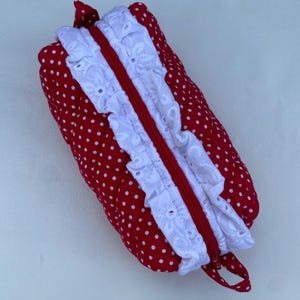May include: Red polka dot zippered pouch with white eyelet lace trim and a red zip. The pouch is rectangular and has a small loop on one end. The fabric is a bright red with white polka dots.