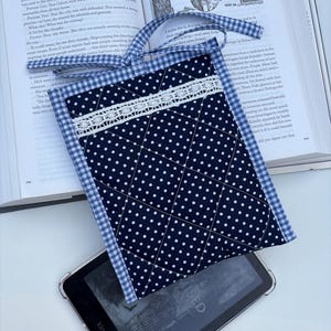May include: A navy blue and white polka dot quilted book sleeve with a white lace accent and a blue gingham border. The sleeve has a blue gingham handle and is resting on a Kindle.