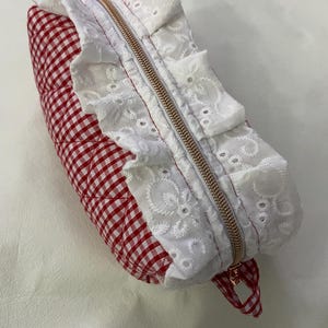 Może przedstawiać: Czerwono-biała pikowana kosmetyczka w kratkę z białym koronkowym wykończeniem i złotym zamkiem błyskawicznym. Torba ma małą czerwoną i białą pętlę w kratkę z boku.