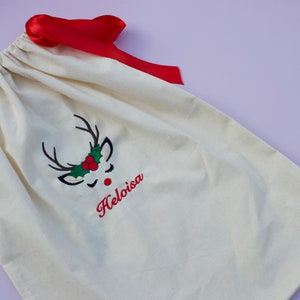 May include: A white canvas gift bag with a red ribbon. The bag has an embroidered reindeer face with a red nose, green holly, and the name "Helloisa" in red.
