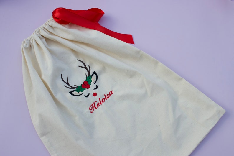 May include: A white canvas gift bag with a red ribbon. The bag has an embroidered reindeer face with a red nose, green holly, and the name "Helloisa" in red.