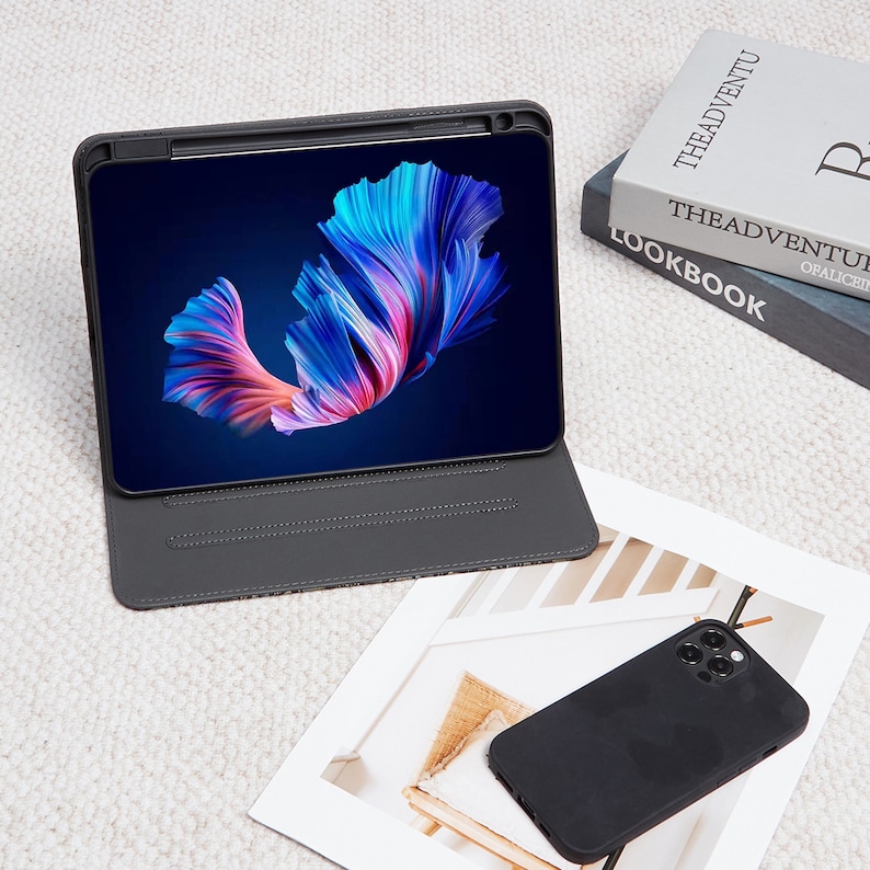 May include: A black leather case with a built-in stand for a tablet. The tablet screen shows a colorful abstract image of a fish. The case is open and the tablet is in a landscape orientation. A black phone case is on a white surface in the foreground.