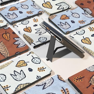以下が含まれることがあります： 茶色、青、白など、さまざまな色の葉の模様が入ったタブレットケースのコレクション。 ケースは白い面に配置され、1つのケースはスタンドで支えられています。 ケースにはステッチされたボーダーがあります。