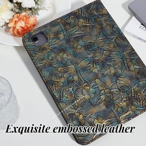 May include: A close-up of a dark green and gold leather iPad case with an embossed floral and dragonfly design. The case has a textured surface and a subtle sheen.