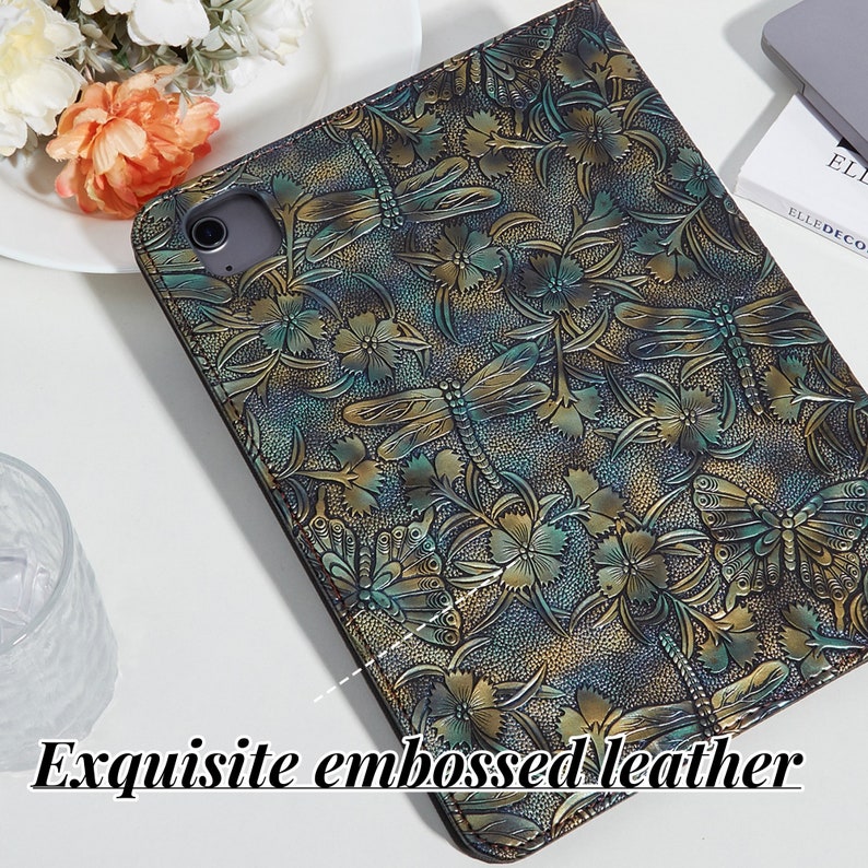May include: A close-up of a dark green and gold leather iPad case with an embossed floral and dragonfly design. The case has a textured surface and a subtle sheen.