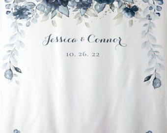 May include: White fabric backdrop with watercolor floral design in shades of blue. The top features a cluster of blue flowers and leaves, with trailing vines. The names "Jessica & Connor" and the date "10. 26. 22" are printed in the center.