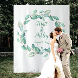 May include: A white wedding banner featuring a watercolour eucalyptus wreath. The names "Matt & Ashley" and the date "August 1. 20yy" are written in script. A couple is kissing in front of the banner.