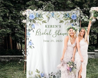 May include: A white backdrop with blue and green watercolor floral designs. The text "Keren's Bridal Shower June 25, 2022" is printed on the backdrop. Two people are standing in front of the backdrop.