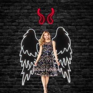 May include: A woman in a floral dress stands before a black brick wall, with a neon angel wing outline and red neon devil horns above. The dress is navy blue with colourful floral embroidery. The neon sign has the text "@ColorfulNeon".