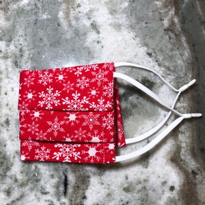 May include: Three red fabric face masks with white snowflake patterns and white elastic ear loops.