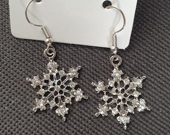 May include: A pair of silver snowflake earrings with clear crystal accents. The earrings are dangle style with a hook closure.
