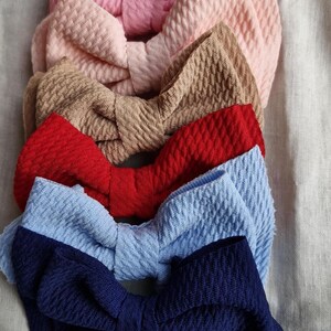 May include: Six fabric hair bows in a row, in shades of pink, beige, brown, red, light blue, and navy. The bows have a textured, ribbed finish. The bows are arranged on a neutral-colored fabric.