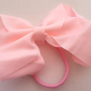 May include: A pink hair tie with a large, pink bow. The bow is made of a textured fabric and has a slightly ruffled appearance.