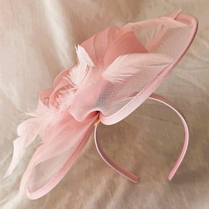 May include: A pale pink fascinator with a headband. The headpiece features a mesh overlay, a large bow, and decorative feathers. The headband is a matching pale pink colour.