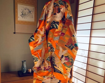 May include: A traditional Japanese wedding kimono with a vibrant orange base and intricate gold embroidery. The kimono features a design of cranes flying over a landscape with stylized clouds and flowers.