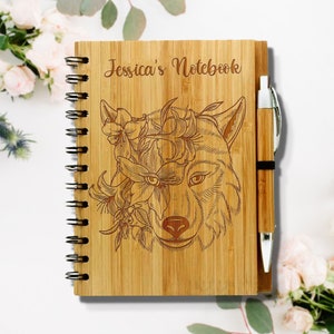 May include: A personalized bamboo notebook with a wolf's head and flowers engraved on the cover. The notebook has a spiral binding and comes with a matching pen. The text "Jessica's Notebook" is engraved on the cover.