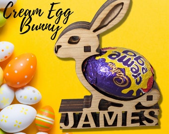May include: Wooden Easter bunny-shaped holder with a purple and gold wrapped chocolate egg. The bunny has cut-out details and the name "JAMES" at the base. Decorative Easter eggs in various colors are scattered around the bunny on a yellow background.