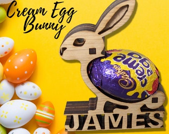 May include: A wooden bunny rabbit cutout holds a purple and gold foil-wrapped chocolate egg. The bunny has a nameplate that reads "JAMIES". The bunny is on a yellow background with several colorful Easter eggs.
