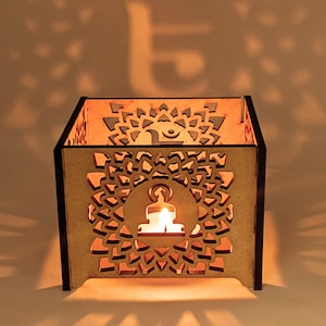 Puede incluir: Un portavelas de madera con un diseño recortado de un Buda meditando y un mandala. El portavelas es marrón y tiene forma cuadrada.