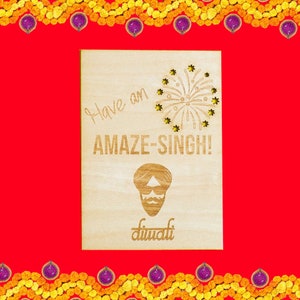May include: A wooden card with a festive message that says "Have an AMAZE-SINGH! Diwali". The card features a cartoon illustration of a man wearing a turban and a mustache. The background is red with a border of yellow flowers.