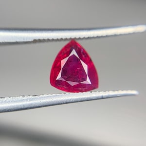 May include: A faceted, triangular ruby gemstone held by tweezers. The ruby is a deep red color with a bright, geometric interior. The tweezers are silver and the background is a soft gray.
