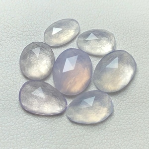 Könnte beinhalten: Sieben schimmernde, facettierte, ovale, hellviolette Edelsteine. Die Steine haben eine durchscheinende, milchige Qualität.