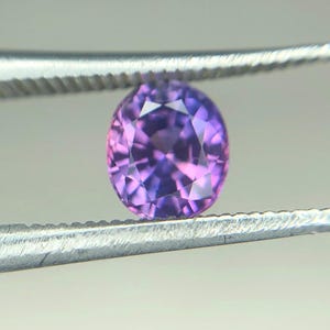 Puede incluir: Una piedra preciosa redonda y facetada con un tono púrpura y rosa. La piedra preciosa está sujeta por unas pinzas.