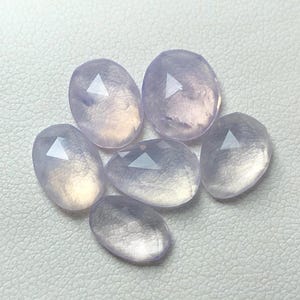 Könnte beinhalten: Sechs facettierte Cabochons aus helllavendelfarbenem Edelstein, mit leicht ovaler Form, liegen auf einer weißen Oberfläche.