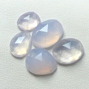 Könnte beinhalten: Fünf blass lavendelfarbene Edelstein-Cabochons mit facettierten Kanten. Die Steine sind durchscheinend und haben einen leichten rosa Farbton.