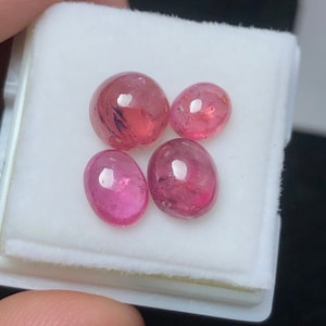 Puede incluir: Cuatro piedras preciosas ovaladas de color rosa en una caja blanca. Las piedras preciosas son lisas y brillantes.
