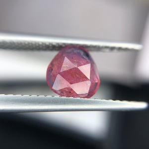 May include: A faceted, pink gemstone with a rose cut. The gemstone is held in tweezers.
