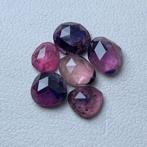 Op de afbeelding: Een assortiment gefacetteerde edelstenen in roze, paarse en violette tinten. De edelstenen hebben verschillende vormen, waaronder ovaal en druppelvormig, en zijn gerangschikt op een wit oppervlak. De edelstenen hebben een gepolijst, reflecterend oppervlak.