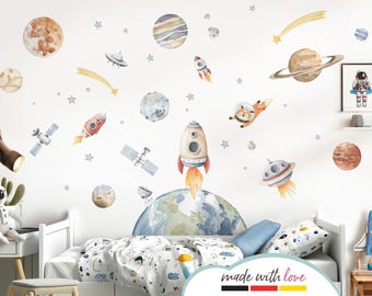May include: A set of space-themed wall decals featuring planets, rockets, stars, a spaceship, and a fox wearing an astronaut helmet. The decals are in shades of blue, red, orange, and yellow. The decals are shown on a white wall above a bed with blue and white bedding.