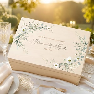 Può includere: Una scatola rettangolare in legno chiaro con un design a ghirlanda floreale stampato nei toni del verde e del bianco. Il testo centrale recita: Hand in hand ein leben lang, Florian & Greta, 18.03.2026. La scatola ha una finitura liscia.